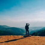 A lone traveler stands on a hilltop, gazing over a vast mountain landscape under a clear blue sky.