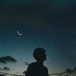 A lone silhouette stands beneath a crescent moon and starry sky, evoking a sense of solitude and wonder.