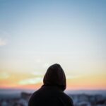 A solitary figure in a hoodie gazes at a vibrant sunset from a rooftop, creating a peaceful silhouette.